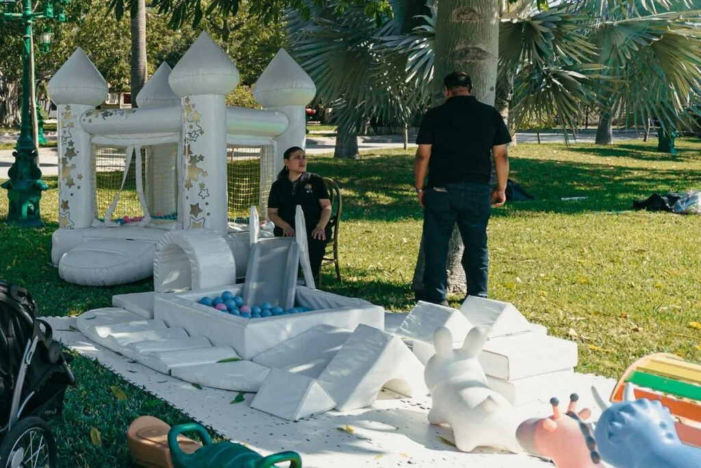 Two crew members beside a white bounce house and soft-play set they have just set up in a park